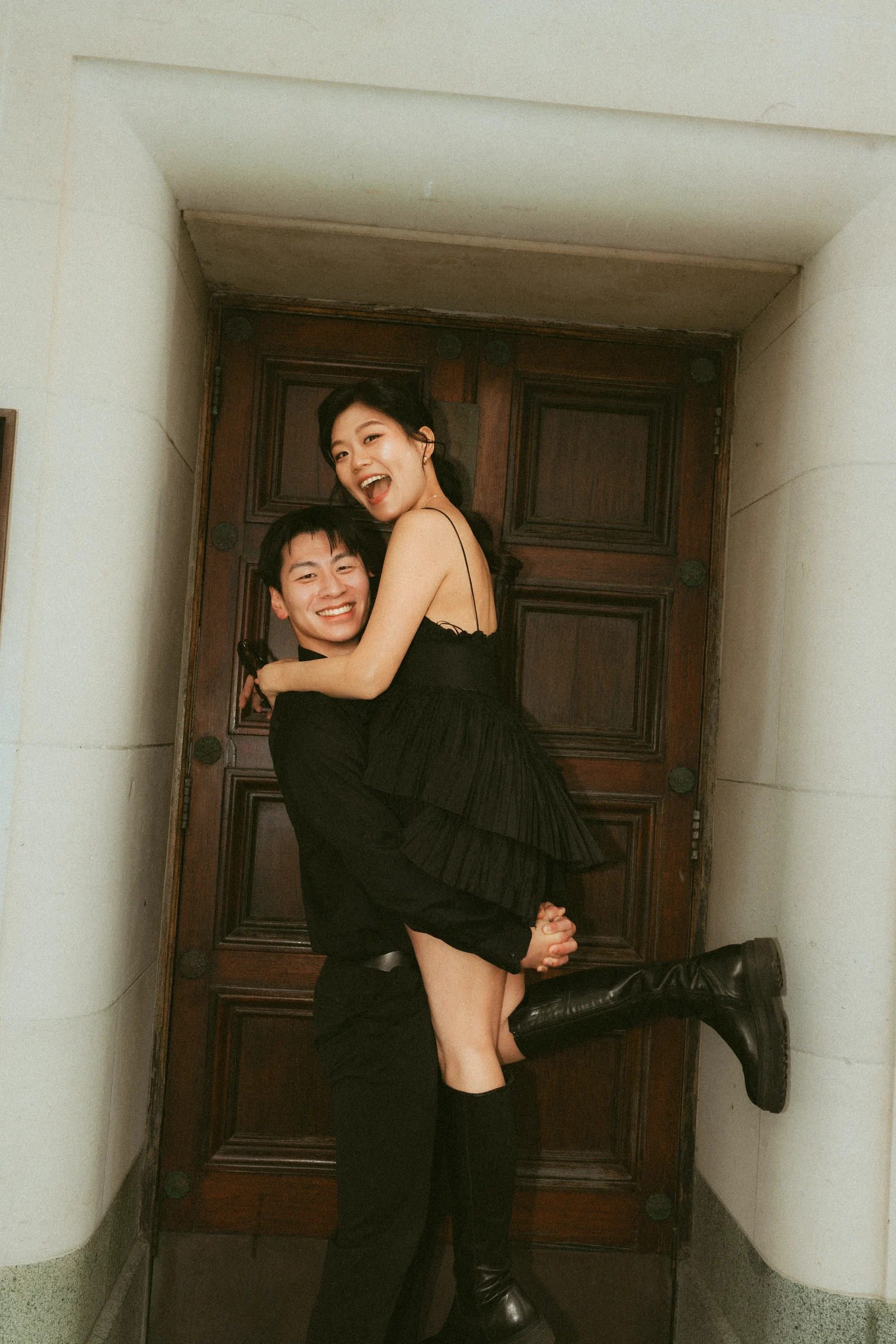Emily being held by Benny in front of a wooden doorway, both wearing black